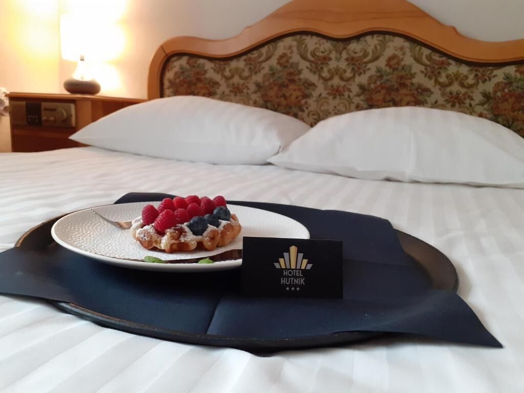 Siististi pedattu sänky, jossa on valkoiset lakanat ja kukka-aiheinen pääty. Tarjottimella on lautanen, jossa on jälkiruokaa marjoineen, ja sen vieressä on käyntikortti, jossa lukee ”HOTEL HUTNIK”.