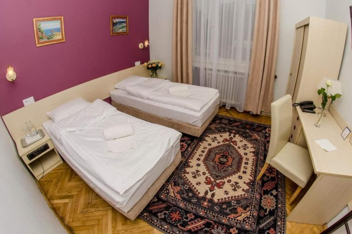 A room with two beds, a desk, a chair, a mirror, a nightstand, and a patterned rug.