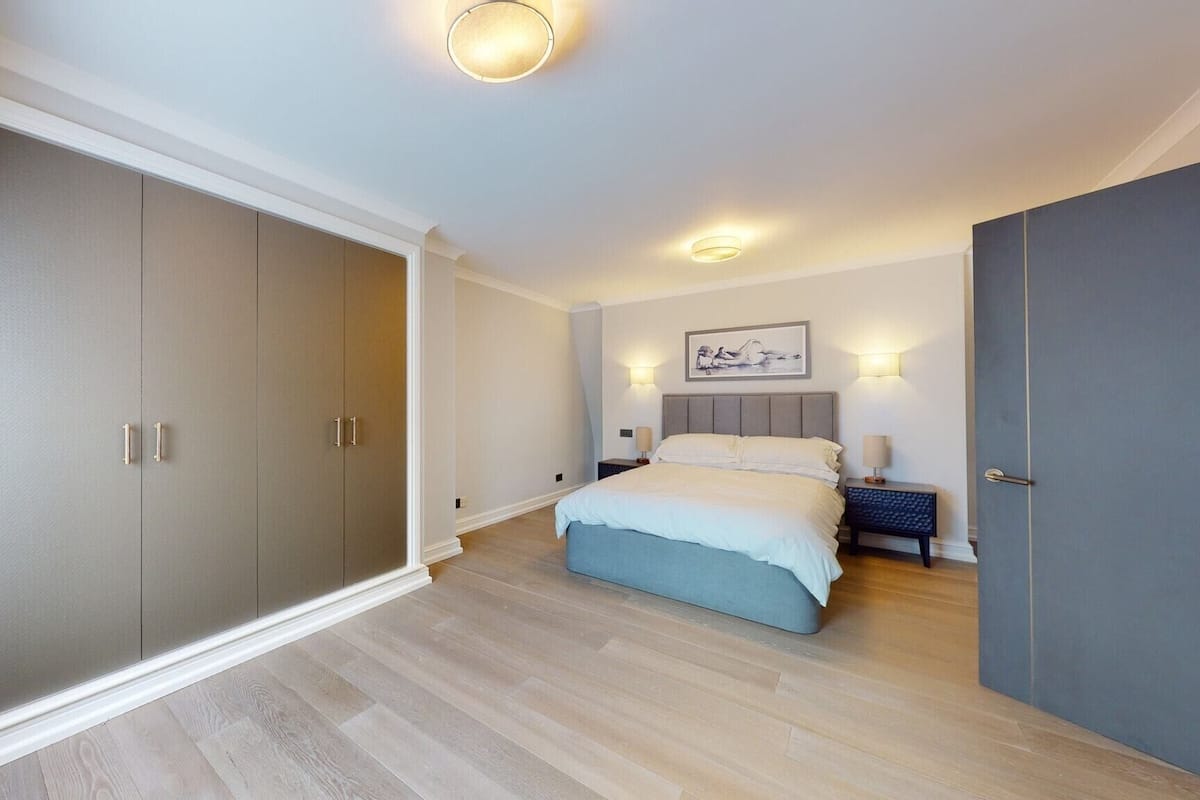 A bedroom with a large wardrobe, a bed with a blue headboard, and two bedside tables with lamps.