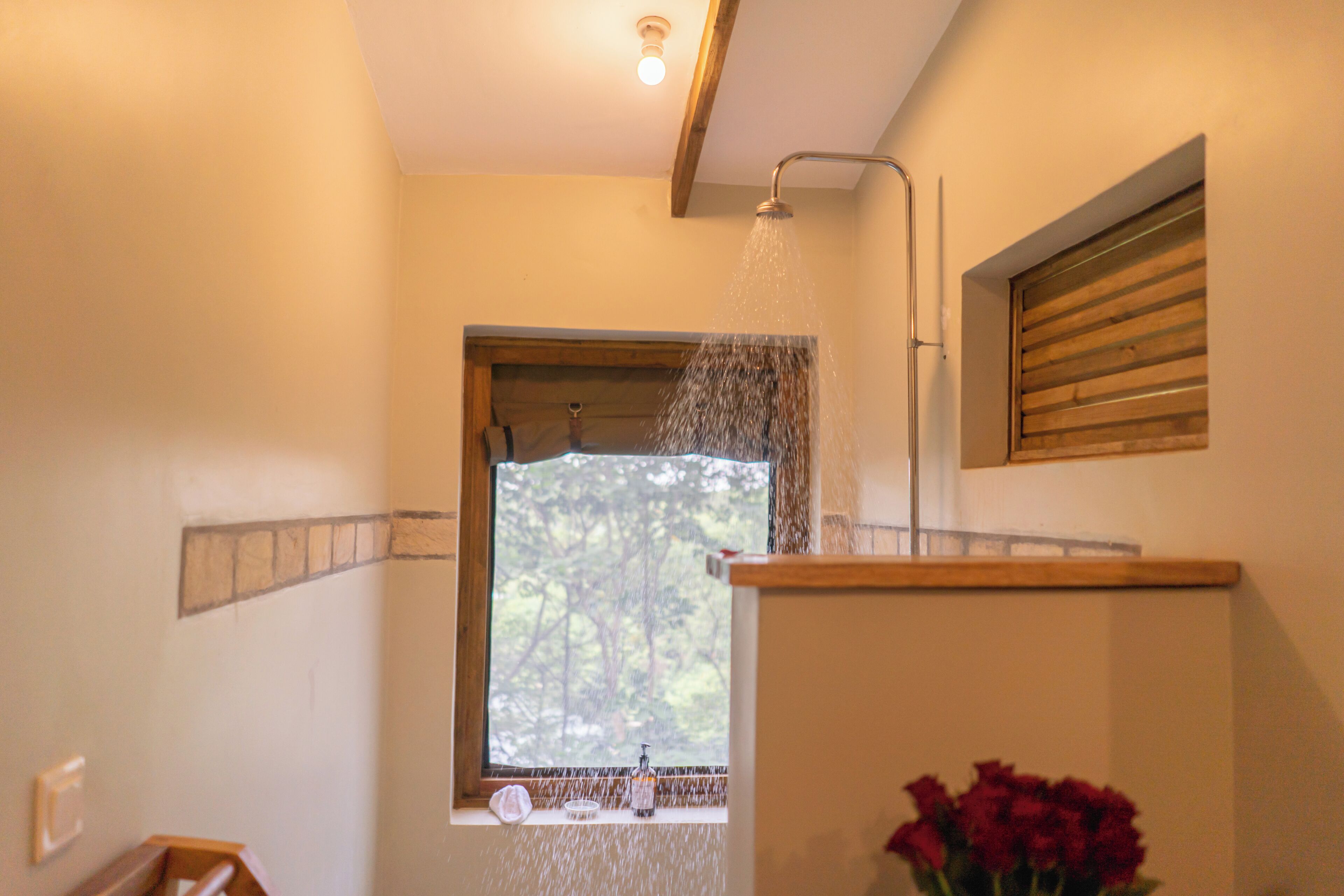 A shower with water running, a window with a view of trees, and a wooden shelf with a bottle and glass.