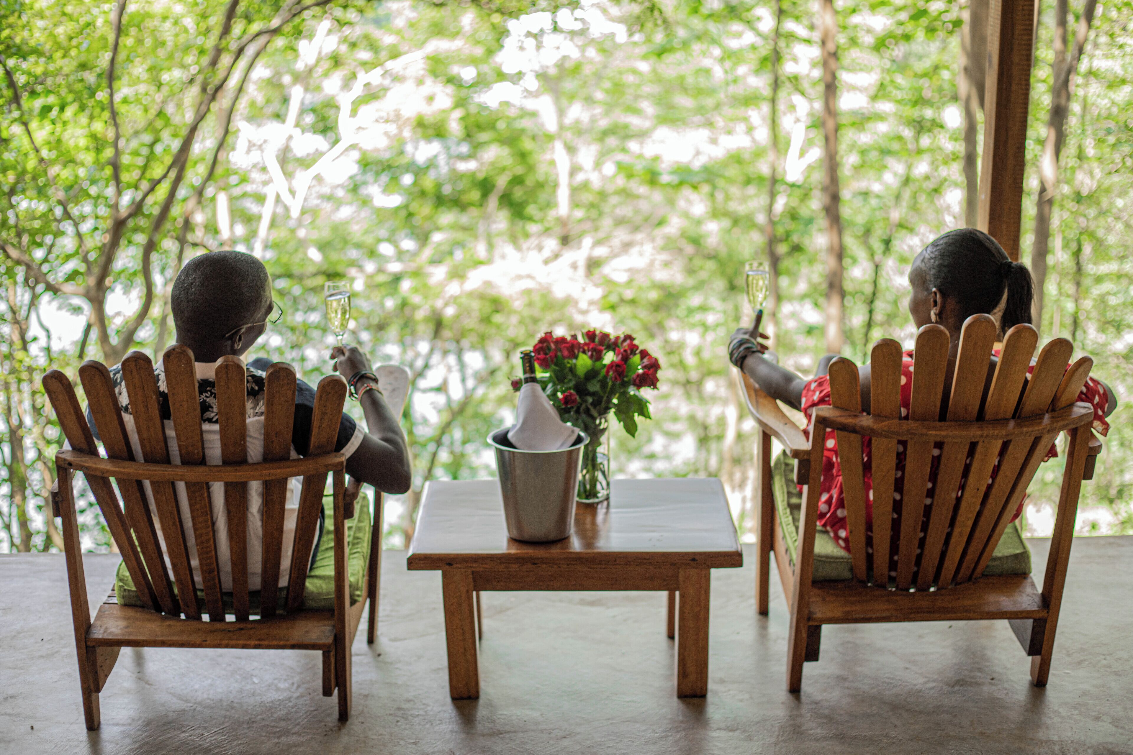 Two wooden chairs facing each other with a small table between them, a vase with red flowers, and a glass of champagne.