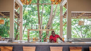 A bar area with wooden chairs, a counter with bottles and glasses, and a view of trees through the structure.