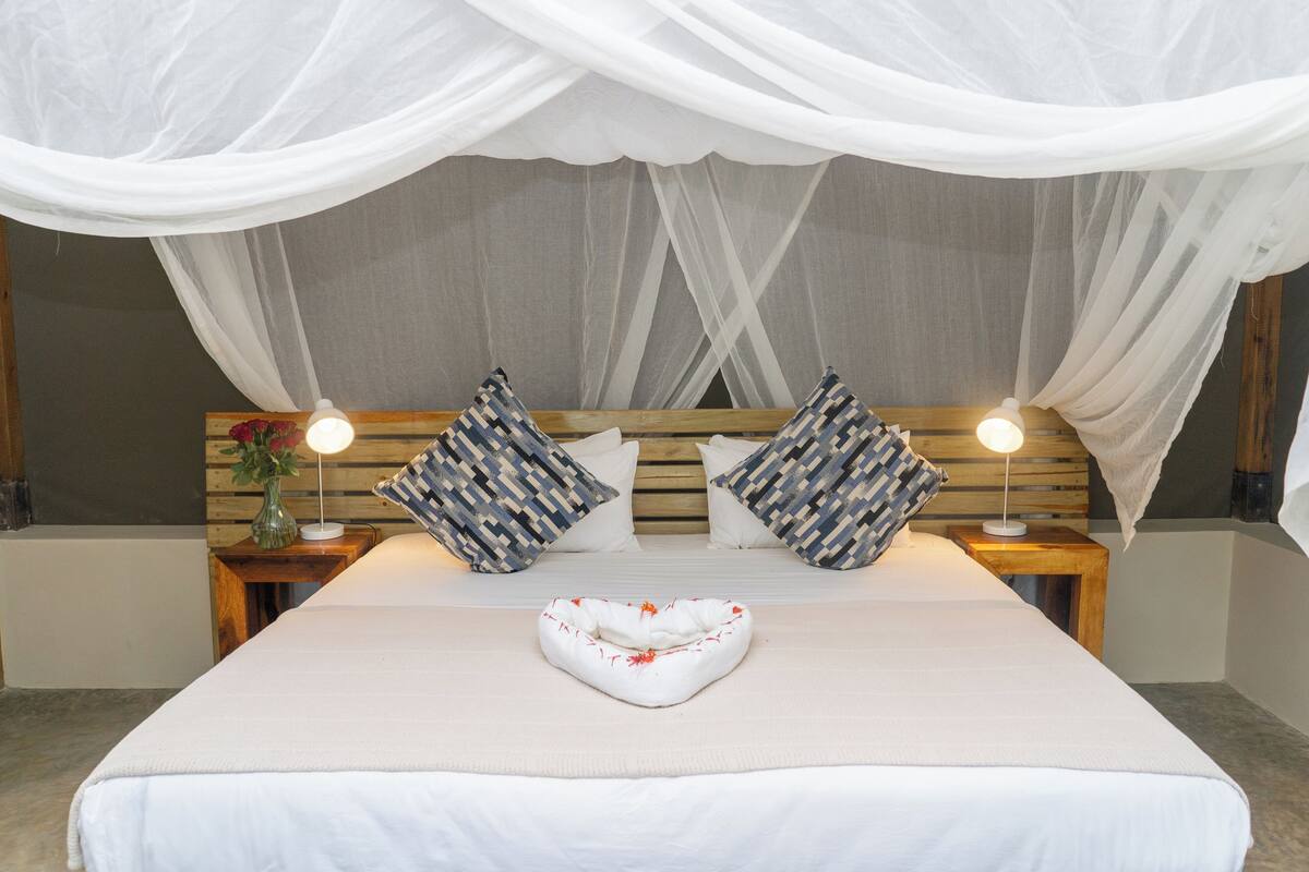 Una cama bien tendida con una colcha blanca, dos almohadas estampadas y una almohada decorativa en el centro. Hay dos mesitas de noche con lámparas y un florero con flores.