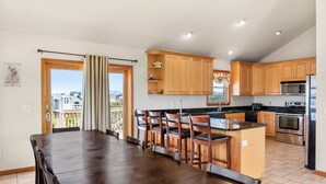 A spacious kitchen with wooden cabinets, a central island with bar stools, a stainless steel refrigerator, and a large window overlooking a balcony. - Bohemian Breeze: Oceanside | Pool and Hot Tub (Southern Shores)