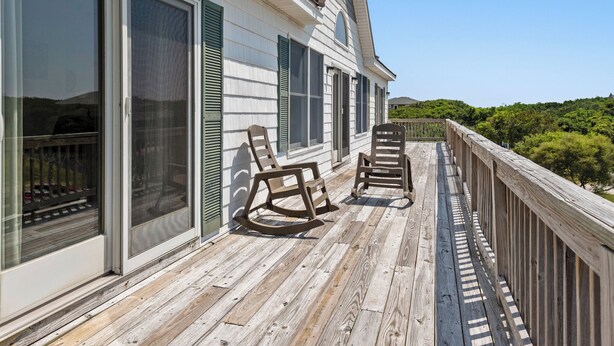 A wooden deck with rocking chairs and a view of a forested hill. - Bohemian Breeze: Oceanside | Pool and Hot Tub (Southern Shores)