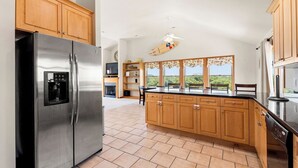 A kitchen with wooden cabinets, a stainless steel refrigerator, a dishwasher, and a window with a view of greenery. - Bohemian Breeze: Oceanside | Pool and Hot Tub (Southern Shores)