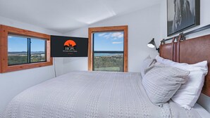 Ein Schlafzimmer mit einem großen Bett, zwei Fenstern mit Holzrahmen, einem Wandfernseher und einem gerahmten Bild an der Wand.