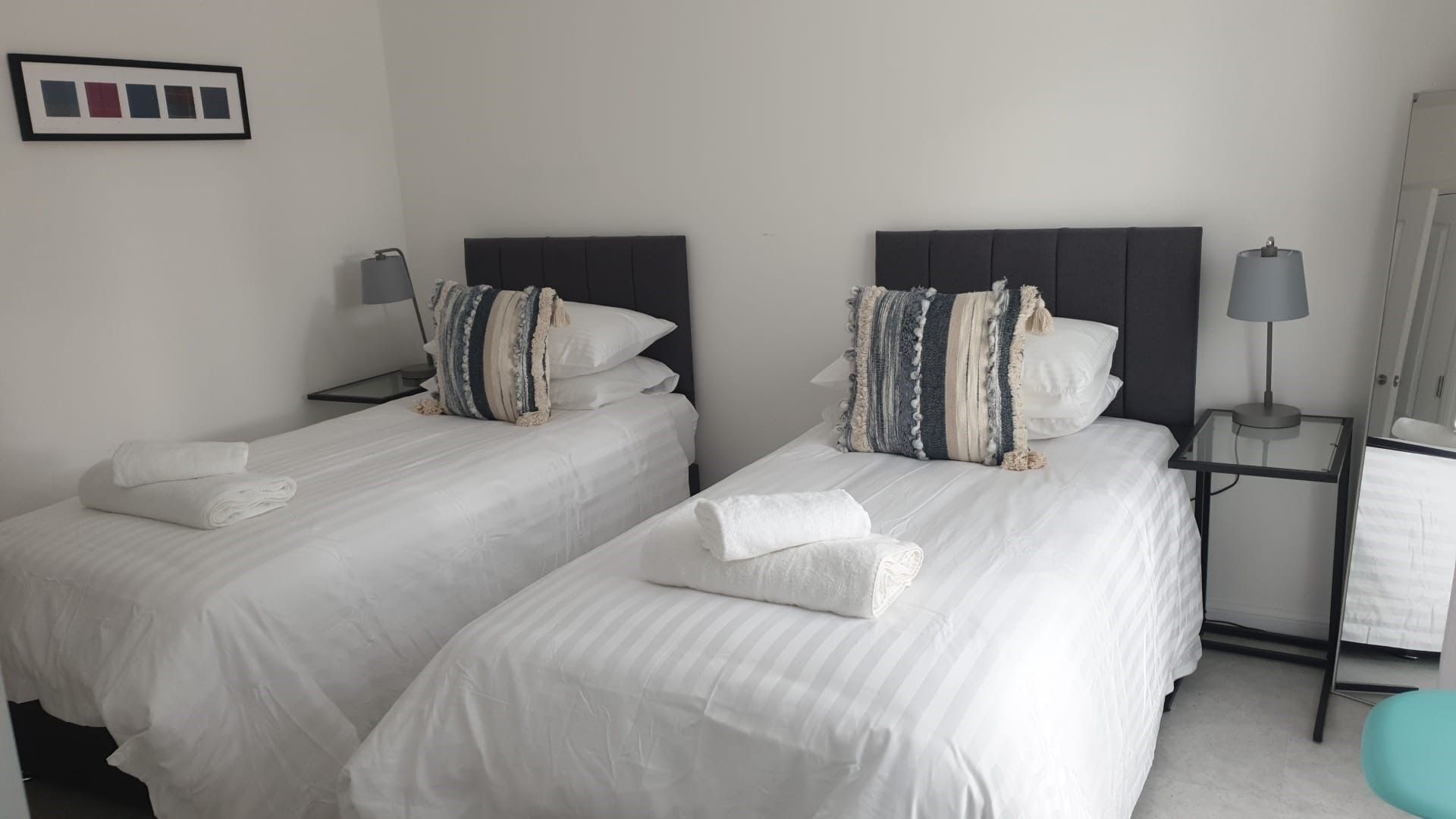 Two single beds with white linens and striped pillows, a small bedside table with a lamp, and a framed picture on the wall.