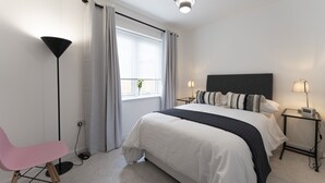 A modern bedroom with a large bed, bedside tables with lamps, a floor lamp, a pink chair, and a window with curtains. - Mhuilinn Apartment  -  an apartment that sleeps 3 guests  in 2 bedrooms (Stirling)