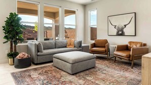 A spacious living room with a grey sectional sofa, two brown leather armchairs, a patterned rug, and a large window offering a view of the outdoors.