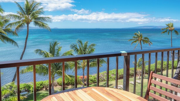 A wooden deck with a table and chairs, overlooking a tropical beach with palm trees and clear blue water.