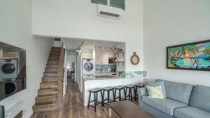 A modern kitchen with a central island, wooden staircase, and a living area with a sofa and bar stools.
