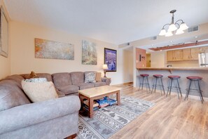 A modern living room with a sectional sofa, a coffee table, and a rug. There are four bar stools and a kitchen area with a refrigerator and cabinets.