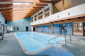 Ein Hallenbad mit blauem Boden, Holzbalken an der Decke und einem großen Fenster auf der rechten Seite.