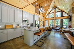 A spacious kitchen with white cabinets, a large island with bar stools, and a stone fireplace. - 2+ Bedroom New Modern cabin Loaded with AMENITIES! SLPS 8 (Broken Bow)