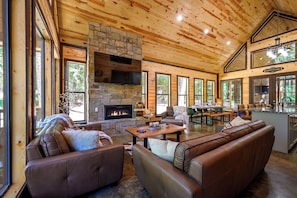 A spacious living area with a stone fireplace, leather sofas, and large windows offering views of the outdoors. - 2+ Bedroom New Modern cabin Loaded with AMENITIES! SLPS 8 (Broken Bow)