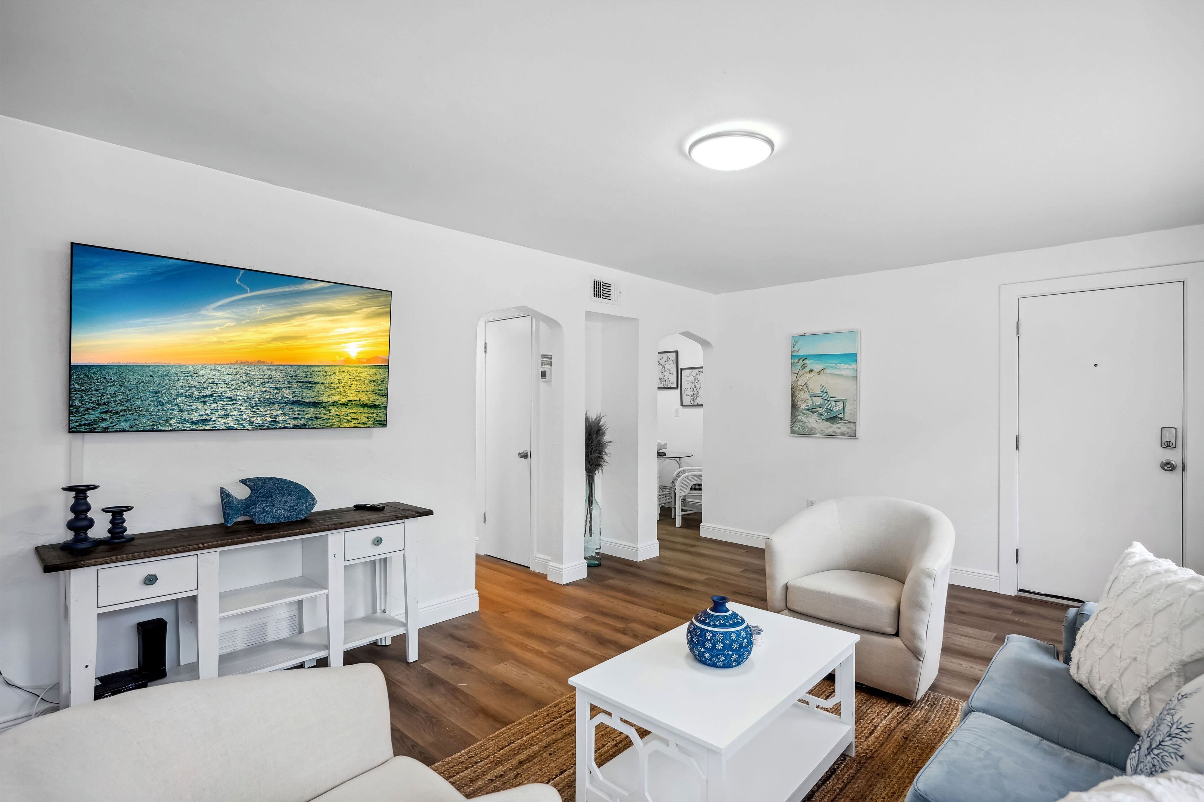 Ein modernes Wohnzimmer mit einem großen Gemälde, das einen Sonnenuntergang über dem Meer zeigt, einem weißen Sofa, einem blauen Sessel und einem weißen Couchtisch.