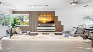 Una sala de estar moderna con un televisor de pantalla plana grande montado en una pared de paneles de madera, un sofá, una mesa de centro y una zona de comedor con una mesa y sillas.