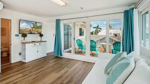 Una habitación moderna con suelo de madera, sofá blanco, cortinas azul verdoso y vistas a un patio exterior con sillas.