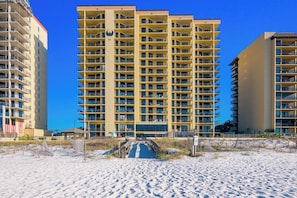 A high-rise hotel with multiple balconies, a central entrance, and a sandy beach in the foreground.