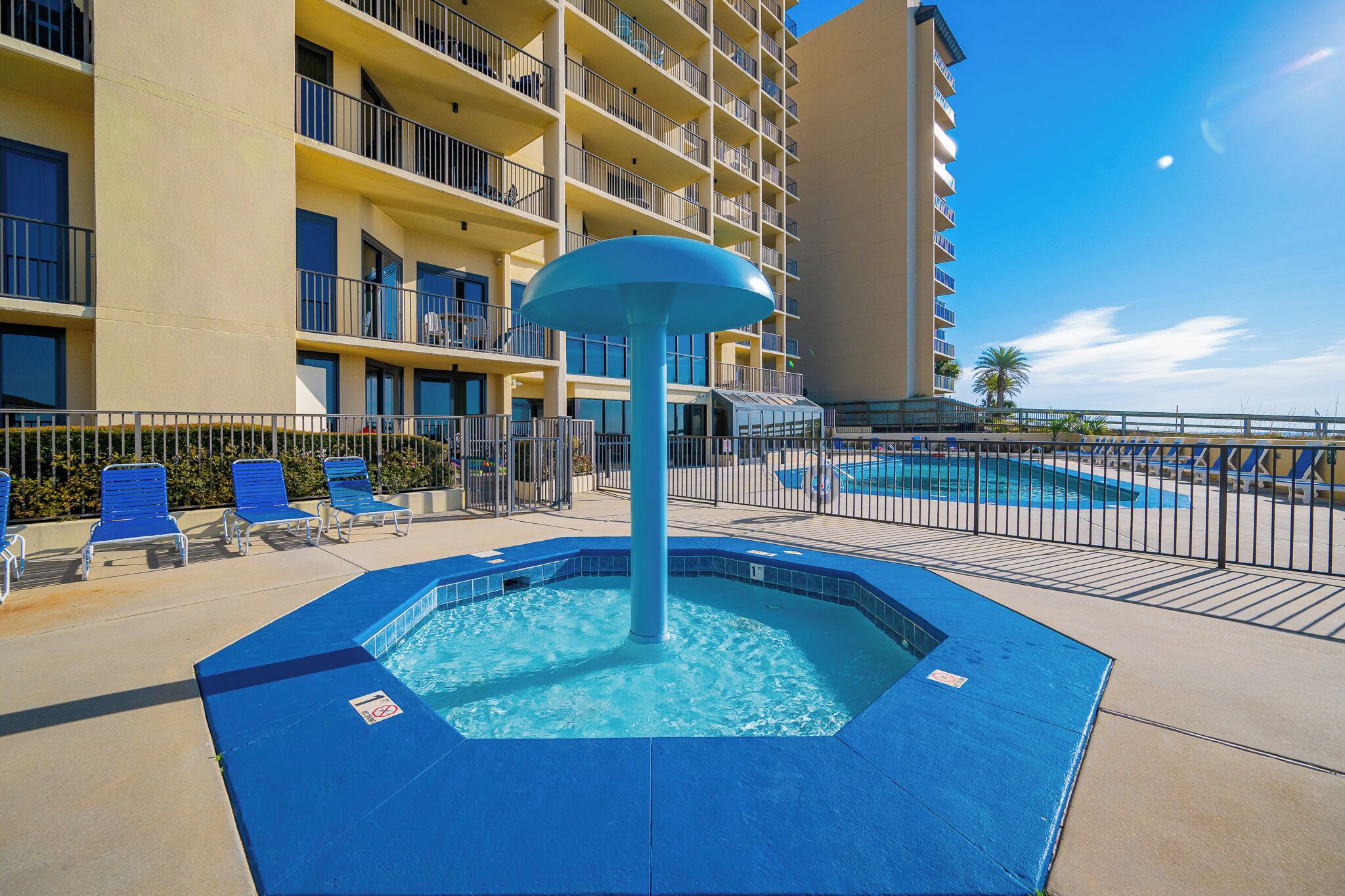 A hotel pool area with blue chairs, a central water feature, and a building with balconies.