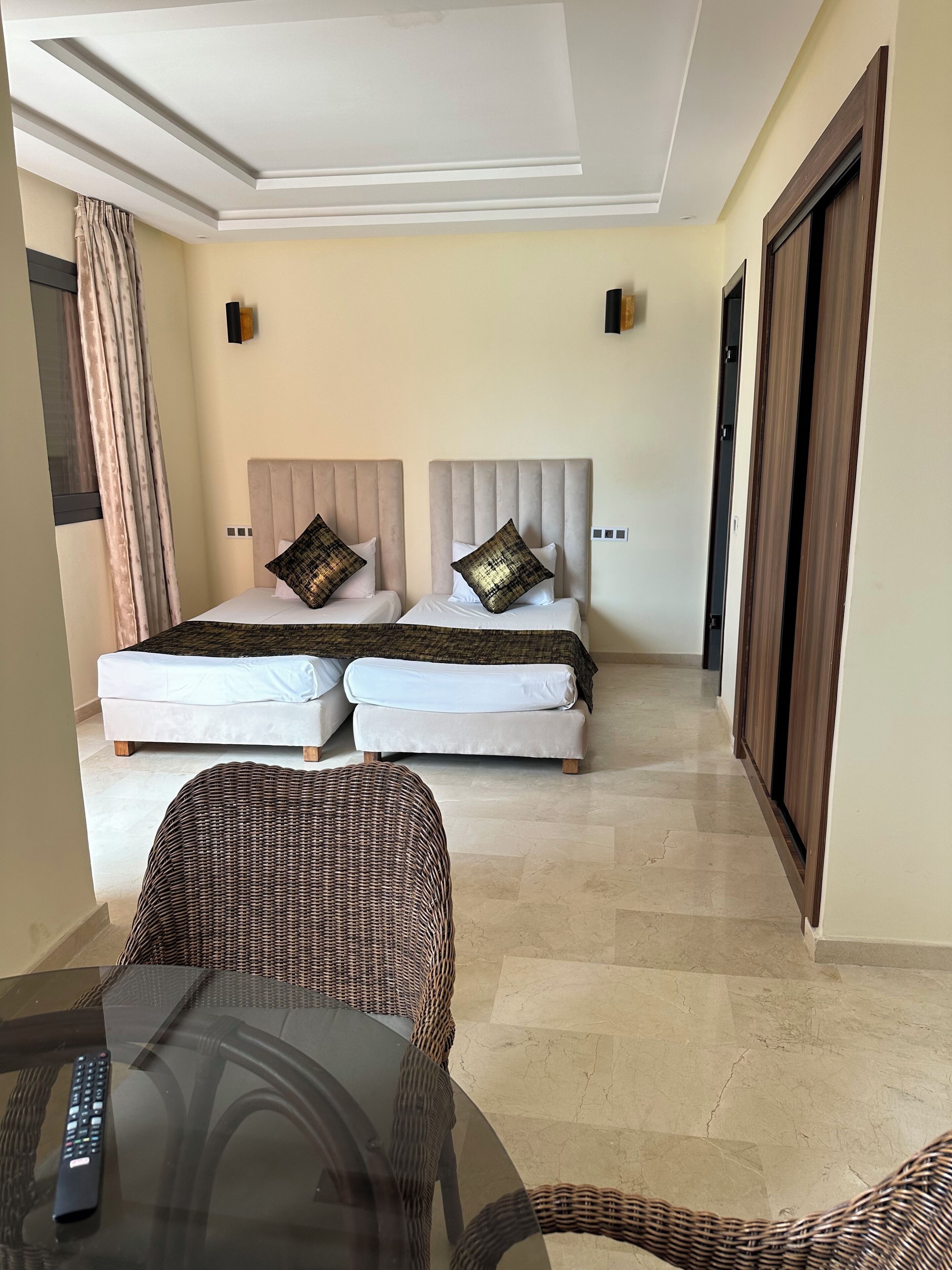 Ein Hotelzimmer mit zwei Betten, einem Korbstuhl, einem Glastisch und einer Fernbedienung.