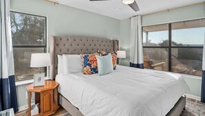 Une chambre avec un grand lit, un ventilateur de plafond et deux fenêtres.