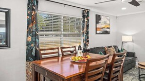 A dining area with a wooden table set for two, a fruit plate, wine glasses, and a bottle. There is a window with blinds, a ceiling fan, and a sofa in the background.
