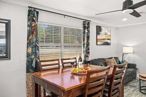 Un comedor con una mesa de madera dispuesta para dos personas, un plato de fruta, copas de vino y una botella. Se aprecia una ventana con persianas, un ventilador de techo y un sofá al fondo.