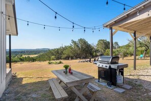 Un pati amb una barbacoa, taula de pícnic i llums penjants, davant d'un fons d'arbres i turons.