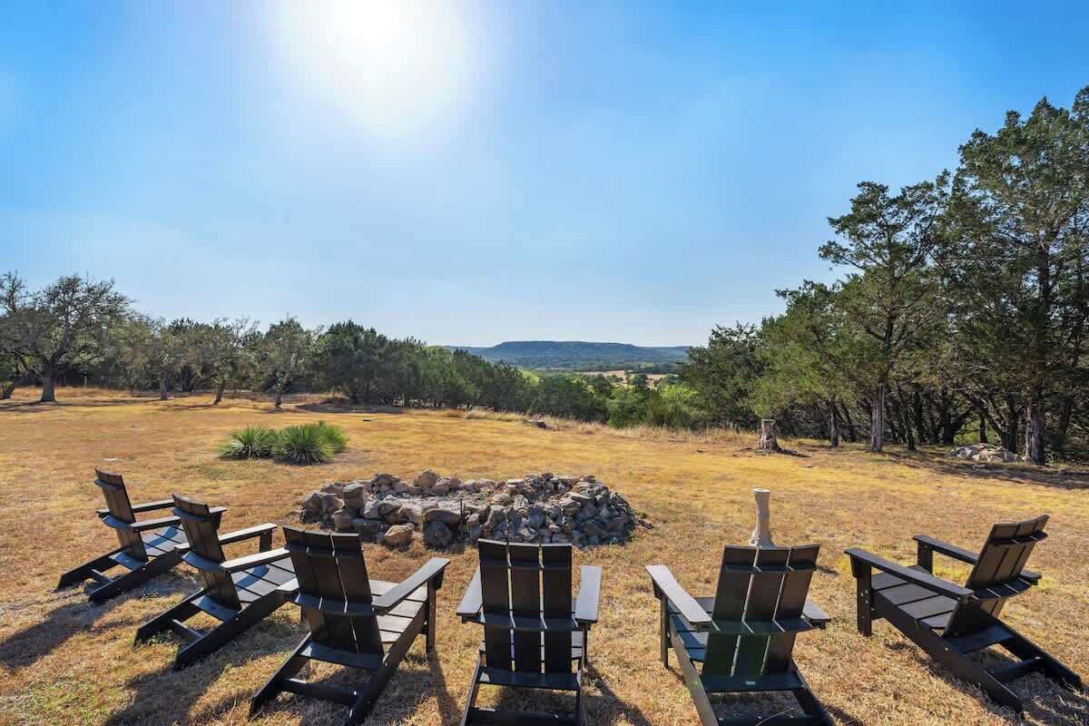 Un camping con un hogar rodeado de sillas Adirondack, con árboles y colinas distantes al fondo.