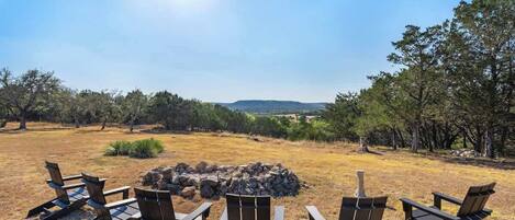 Un camping con un hogar rodeado de sillas Adirondack, con árboles y colinas distantes al fondo.