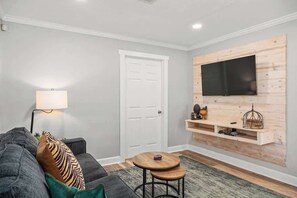 Un salon moderne avec un canapé gris, une petite table ronde, un téléviseur fixé au mur et une porte blanche.