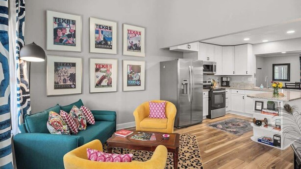 A modern living room with a teal sofa, a yellow armchair, and a coffee table. There are framed pictures on the wall, a refrigerator, and a microwave in the kitchen area.