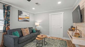 A modern living room with a grey sofa, a round wooden table, a lamp, and a television on a wooden shelf.