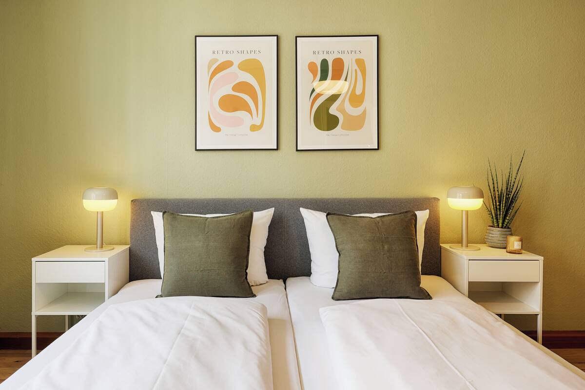 Un lit double avec une literie blanche, deux tables de chevet avec des lampes et deux œuvres d’art abstraites encadrées accrochées au mur.