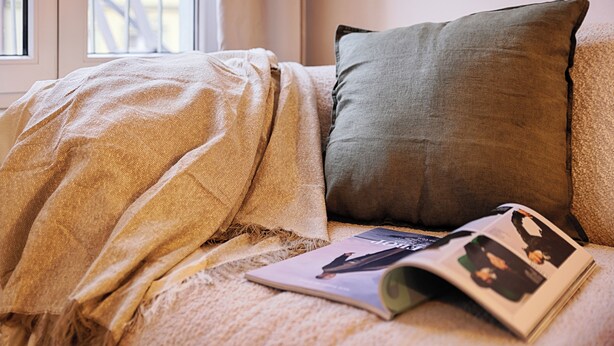 A bed with a beige blanket and a green pillow, a magazine on the bed, and a window in the background. - homie stay karmeliten (Würzburg)