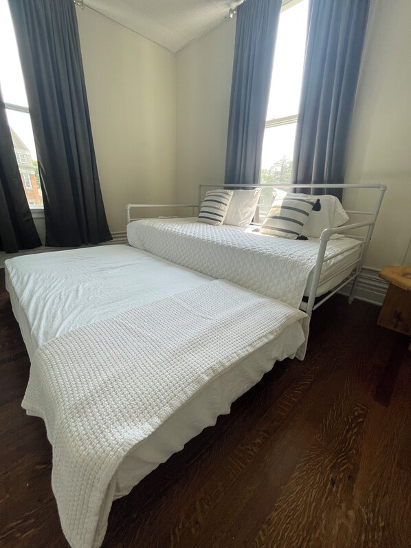 A neatly made bed with a white quilt and pillows, a wooden floor, and a window with curtains. - The Wheat House on Main (Unit 4, 2nd story)) (Geneva)
