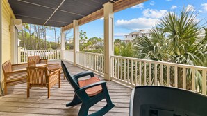 Un espacio al aire libre techado con mobiliario de madera, barandilla blanca y vistas a palmeras y casas.