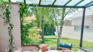 An outdoor dining area with a table set for four, surrounded by chairs, under a glass roof with a view of a playground and greenery. - Maison Familiale de 240m2 Proche Paris (Groslay)
