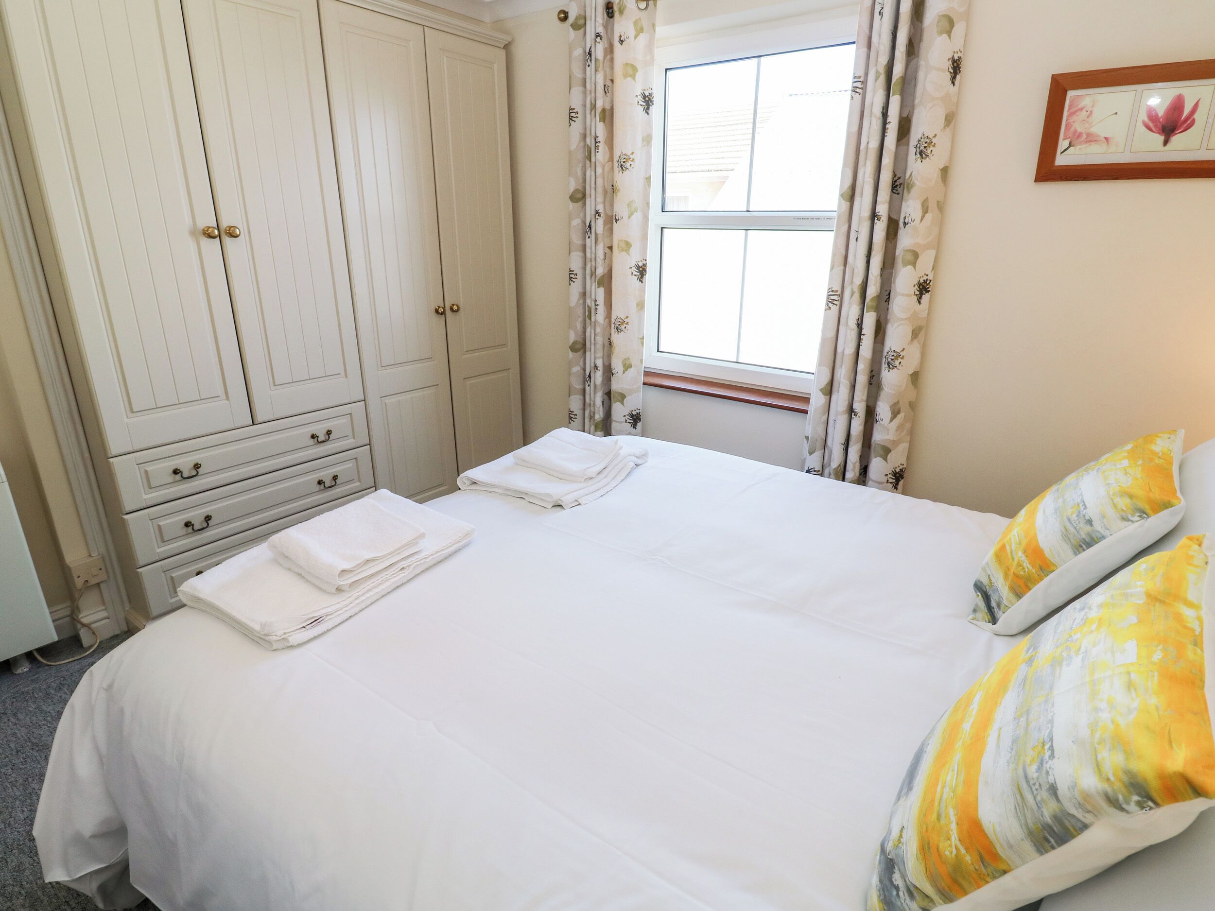 A bedroom with a bed, two pillows, a window with curtains, a wardrobe, and a framed picture on the wall.