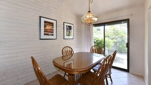 Un coin repas avec une table ronde en bois et six chaises, un mur en briques blanches et une grande fenêtre donnant sur un paysage verdoyant.
