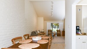 Un coin repas avec une table et des chaises en bois, un mur en briques blanches et une cuisine visible au fond.