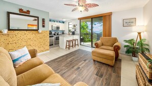 A spacious living area with a sofa, a chair, a kitchen in the background, and a balcony with a view.