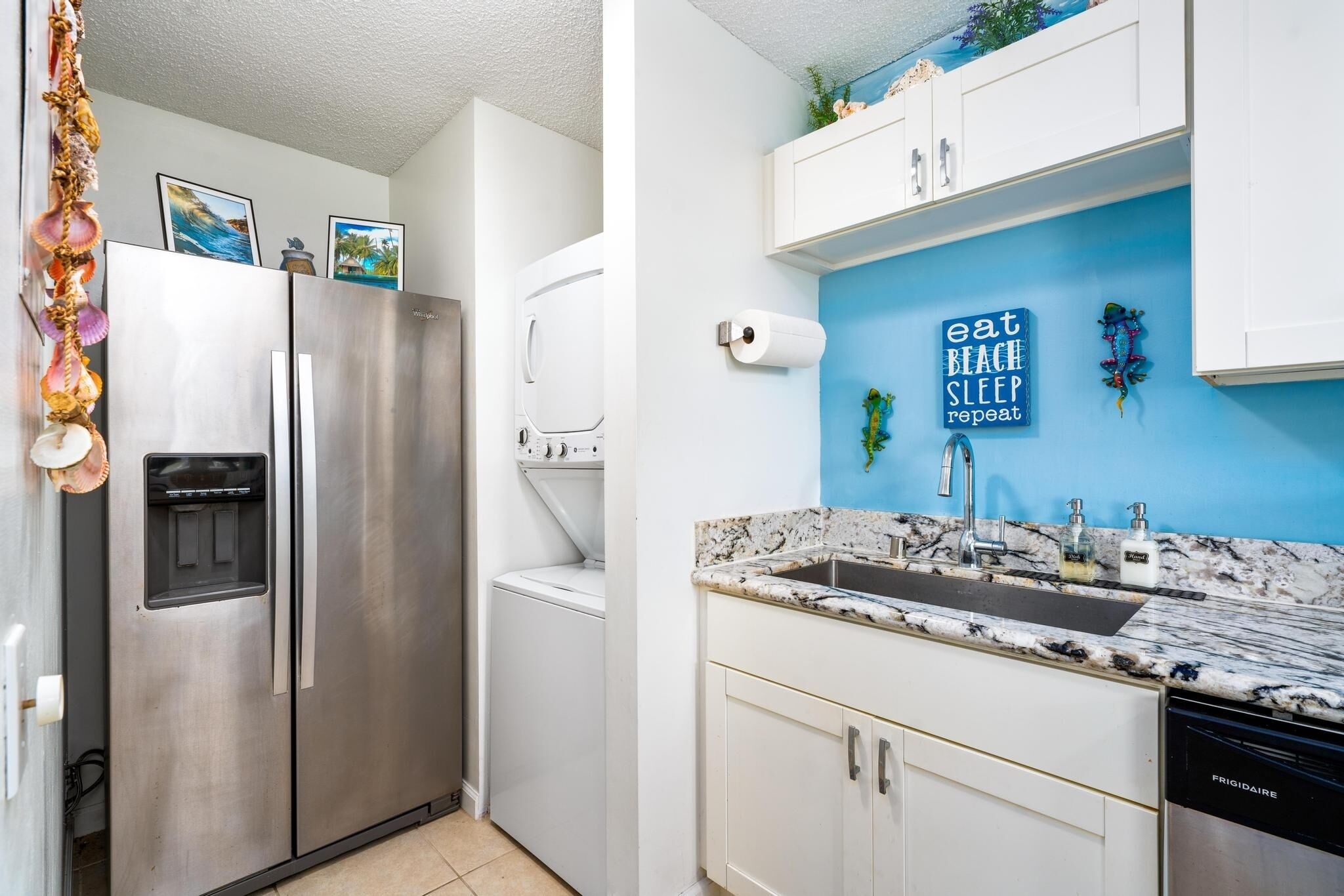 A kitchen with a stainless steel refrigerator, a built-in washer and dryer, a sink with a marble countertop, and a blue wall with a motivational quote.