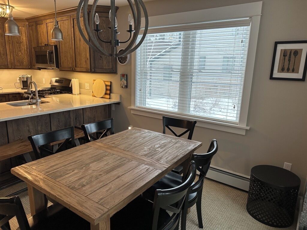 A kitchen with wooden cabinets, a dining table, chairs, a black trash can, and a window with blinds.
