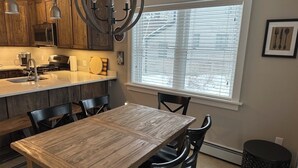 Una cocina con armarios de madera, una mesa de comedor, sillas, un cesto de basura negro y una ventana con persianas.
