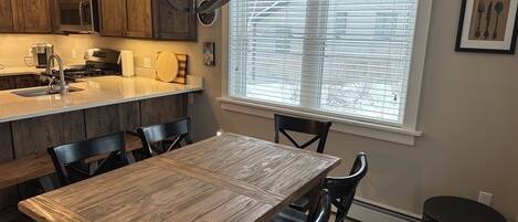 A kitchen with wooden cabinets, a dining table, chairs, a black trash can, and a window with blinds.