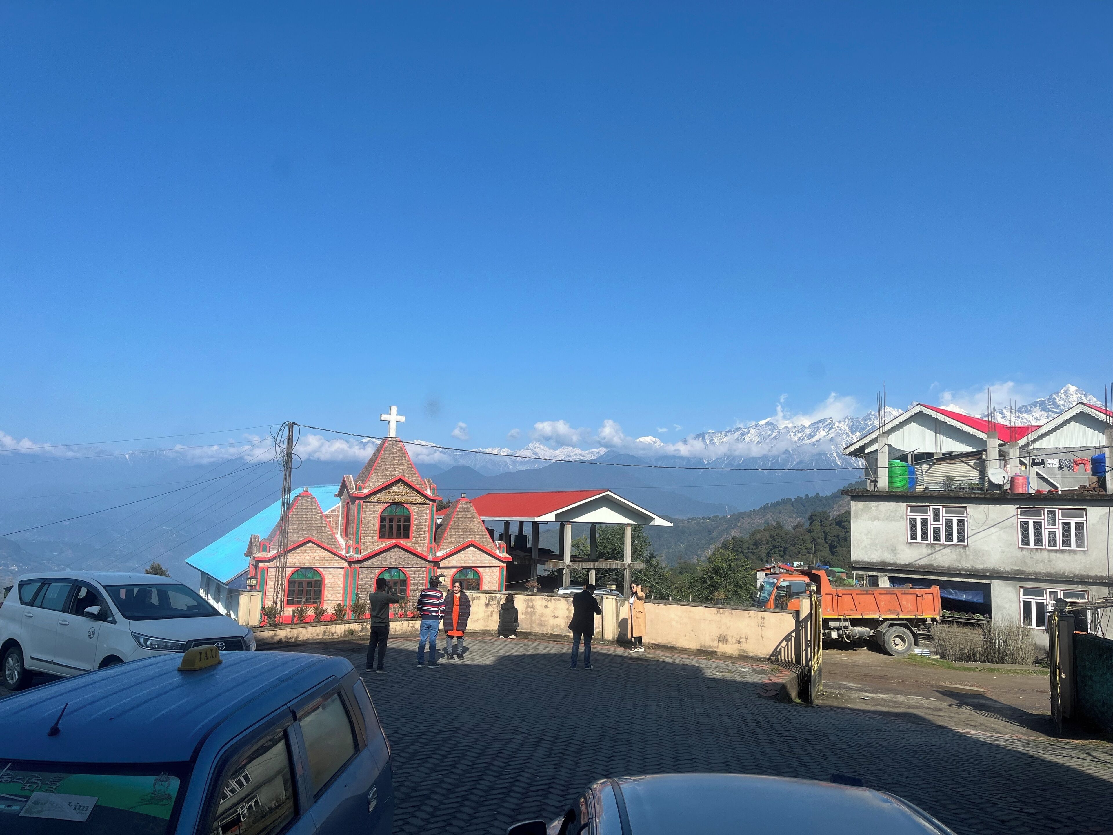A church with a red roof and a cross, surrounded by buildings and a mountainous backdrop.