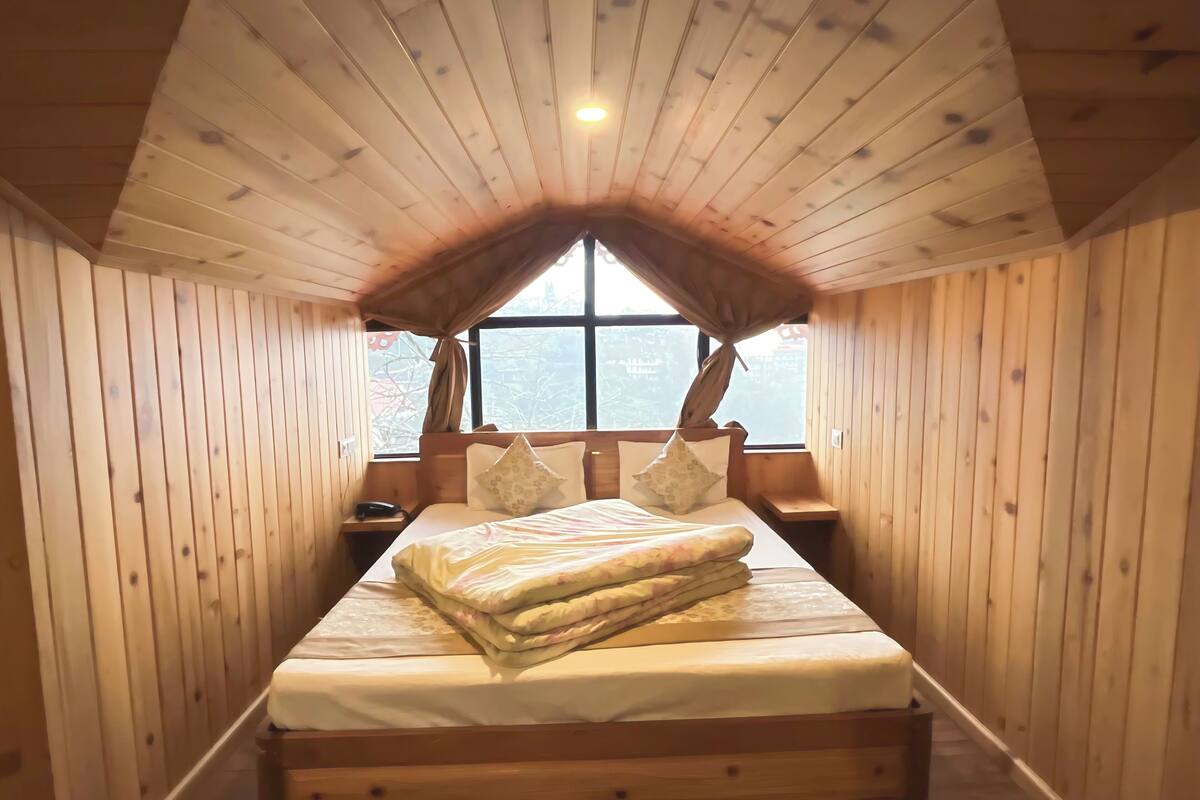 A cozy wooden cabin room with a sloped ceiling, a large bed with pillows, a bedside table, and a window with a view.